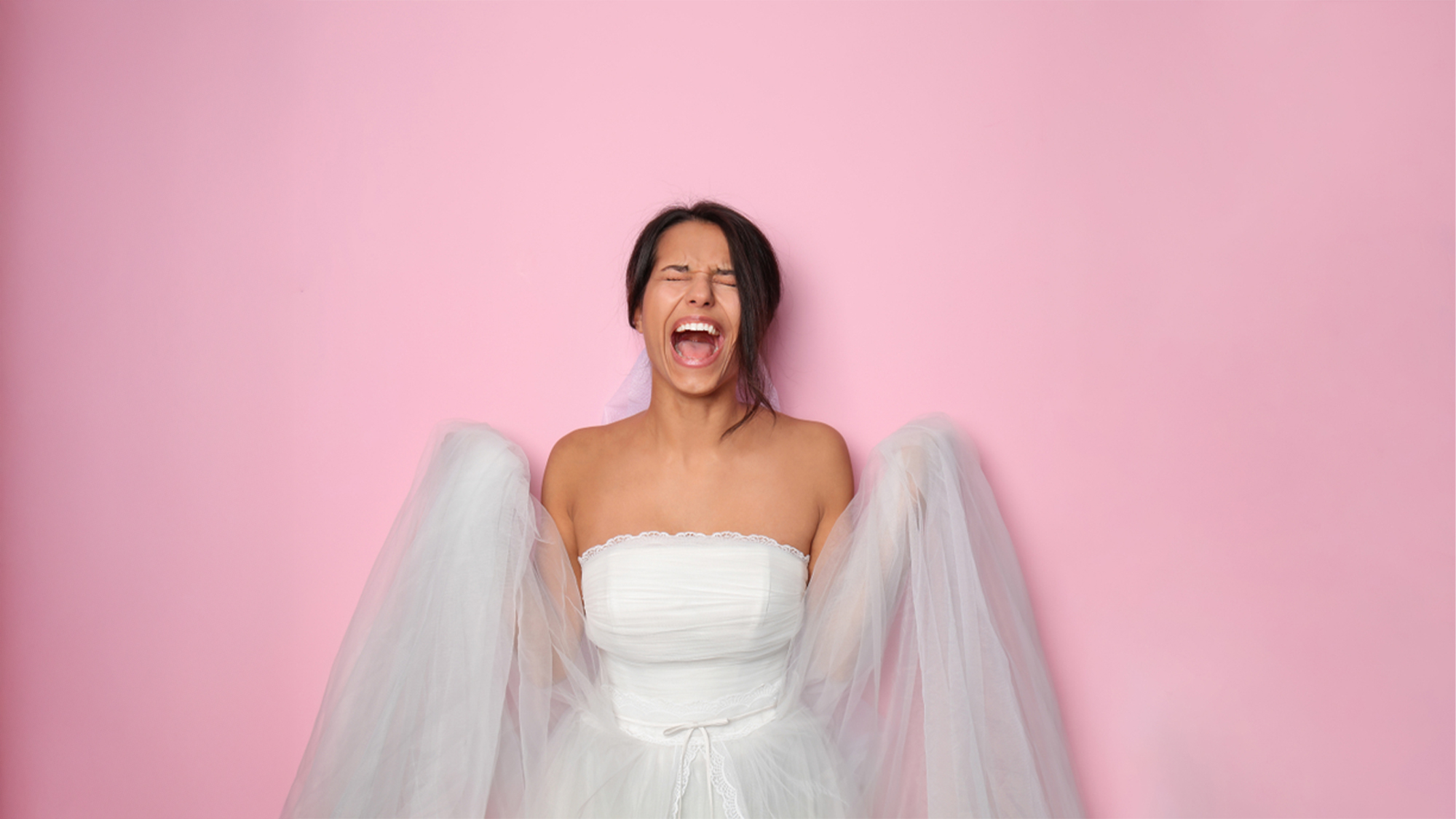 A panicked bride in her white strapless wedding gown and veil screams against a pink background suggesting last-minute dress adjustments