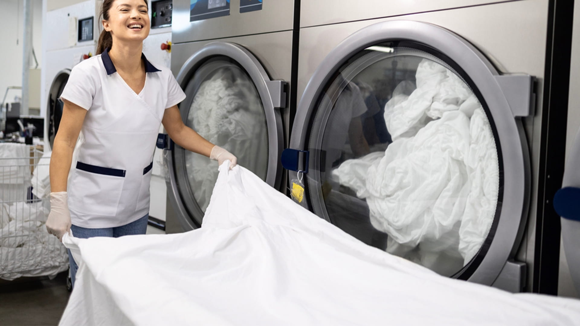 A smiling laundry professional wearing a uniform and gloves pulls large white linens from a commercial washing machine in an industrial facility.